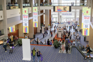 Eine große Menschenmenge bewegt sich durch ein großes Gebäude mit Bannern, Topfpflanzen, Rolltreppen, Geländern, Säulen, Texttafeln und Glastüren, einige tragen Taschen bei der Nationalen Buchmesse.