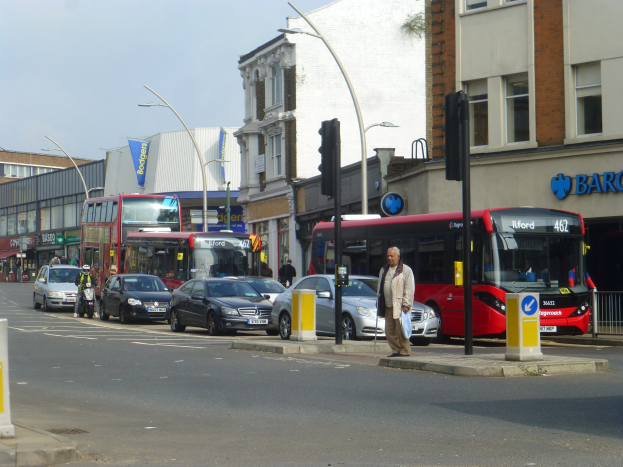 Ein roter Doppeldeckerbus fährt auf einer von hohen Gebäuden gesäumten Stadtstraße, umgeben von Fußgängern, einem Fahrradfahrer, Ampeln, Laternen und Texttafeln unter einem bewölkten Himmel.
