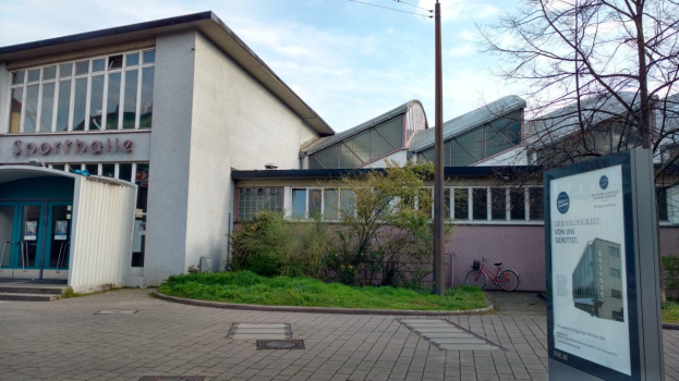 Ein großes Gebäude mit einem Schild davor, umgeben von Bäumen, Pflanzen, Gras, Fahrrädern und einem Pfahl, mit einem bewölkten Himmel im Hintergrund, identifiziert als die Sporthalle, die 2020 eröffnet werden soll.