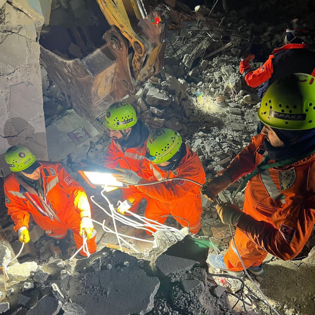 Eine Gruppe von Rettungskräften in orangefarbenen Uniformen und grünen Helmen beseitigt Trümmmer von einem eingestürzten Gebäude mit einem Kran im Hintergrund.
