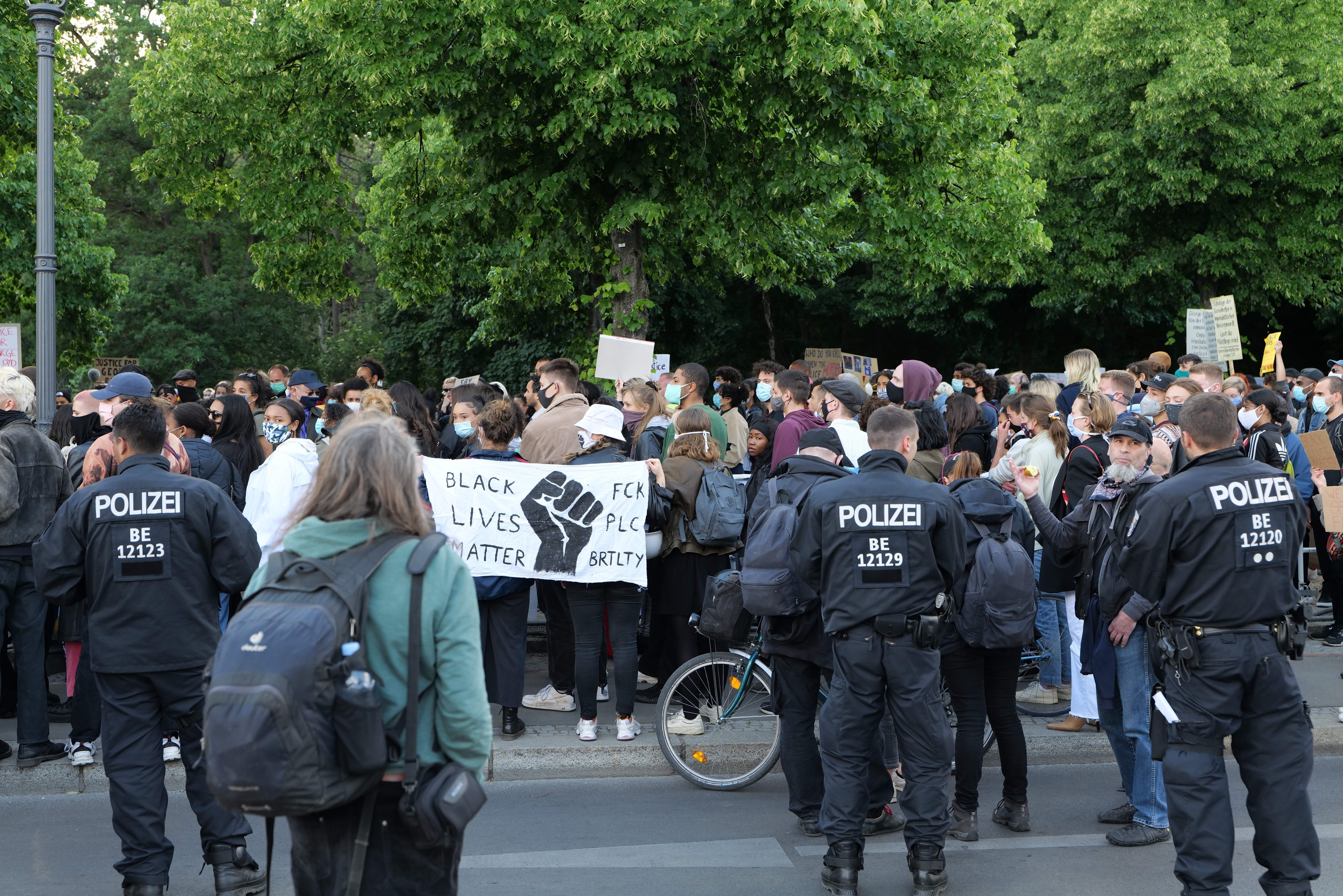 Große Gruppe von Menschen bei einer Black Lives Matter Demonstration in Berlin mit Schildern, einigen mit Mützen und Taschen, vor einem Fahrrad und Bäumen und einem Mast im Hintergrund.