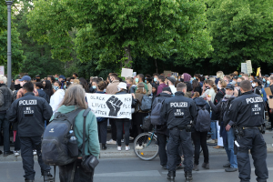 Große Gruppe von Menschen bei einer Black Lives Matter Demonstration in Berlin mit Schildern, einigen mit Mützen und Taschen, vor einem Fahrrad und Bäumen und einem Mast im Hintergrund.