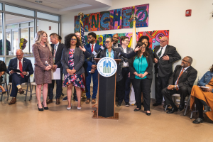 Eine Gruppe von Menschen steht vor einem Podium mit einem Mikrofon und einer Tafel mit dem Text "Sancho-Stadtrat tritt dem Stadtrat bei", während andere auf Stühlen sitzen oder in der Nähe stehen; Flaggen, ein Feuerlöscher und Deckenleuchten sind an der Rückwand zu sehen.