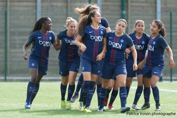 Eine Gruppe junger Frauen, die auf einem Rasenfußballfeld mit Maschendrahtzaun und einer Wand im Hintergrund spielen, mit dem Text "Paris Saint-Germain Women's Soccer" in der unteren rechten Ecke.