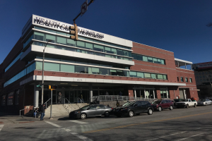 Ein großes Gebäude, das als New York City Health Care Homeless Center identifiziert wurde, mit parkenden Autos davor, Straßenlaternen, Verkehrspolen, Verkehrssignalen, Schildern, Menschen auf dem Gehweg, Geländern, Treppen, Bäumen und einem klaren blauen Himmel.