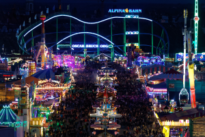 Eine große Menschenmenge steht um einen hell erleuchteten Karneval in der Nacht herum, umgeben von Vergnügungsgeräten, Pfählen, Fahnen und Namensschildern, mit einem dunklen Hintergrund.