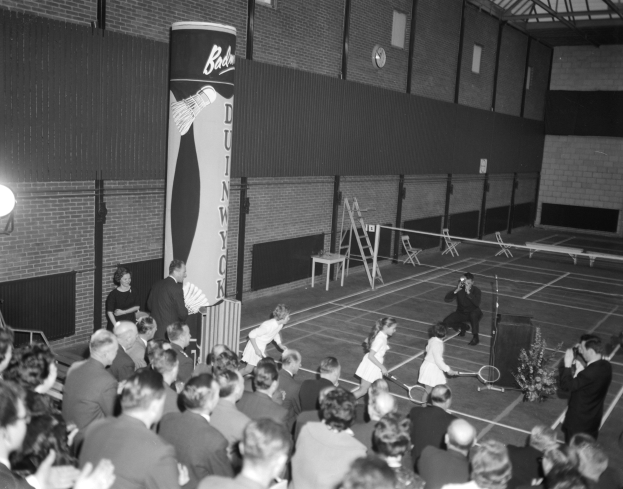 Schwarz-weißes Foto von Menschen, die Tennis in einer Turnhalle spielen, einige stehen und einige sitzen mit Schlägern, eine Uhr an der Wand im Hintergrund.