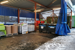 Ein großes Gebäude auf dem Valaisimet Abfallmanagementgelände voller Müll, umgeben von Containern und Fässern, mit Schnee auf dem Boden, Lampen an der Decke und Bäumen, Fahrzeugen und einem klaren blauen Himmel im Hintergrund.