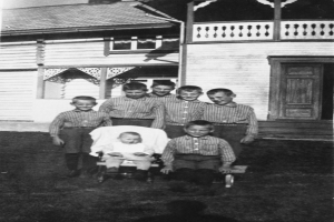 Ein Schwarz-Weiß-Foto einer Gruppe von Kindern, die vor einem Haus stehen, mit einem Baby im Kinderwagen in der Mitte.