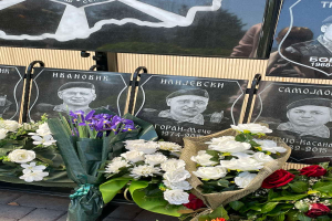 Ein Strauß aus farbenfrohen Blumen liegt auf einem Gehweg neben einem Schild, mit mehreren Gedenktafeln im Hintergrund.