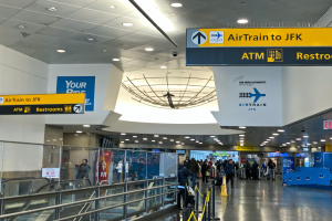 Eine Gruppe von Menschen geht durch eine Flughafenhalle mit einer Rolltreppe auf der linken Seite, Schilder mit Text an den Wänden, Lampen an der Decke und ein paar Menschen, die auf dem Boden stehen, ein Schild liest "Airtrain nach JFK".