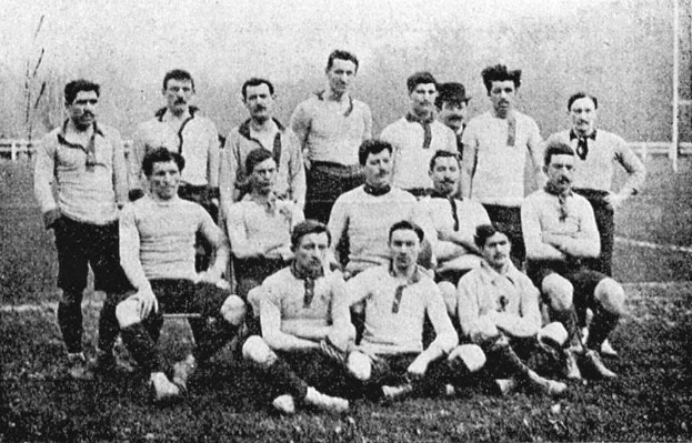 Schwarzes und weißes Foto einer Fußballmannschaft aus den 1920er Jahren mit stehenden und sitzenden Mitgliedern vor Pfählen, Bäumen und einem klaren Himmel.