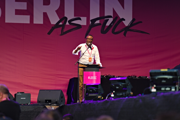 Mann auf einer Bühne vor Publikum stehend, weißes Hemd und braune Hose tragend, Mikrofon haltend, mit einem pinken Banner 'Berlin as Fuck' im Hintergrund und Lautsprechern auf der Bühne.