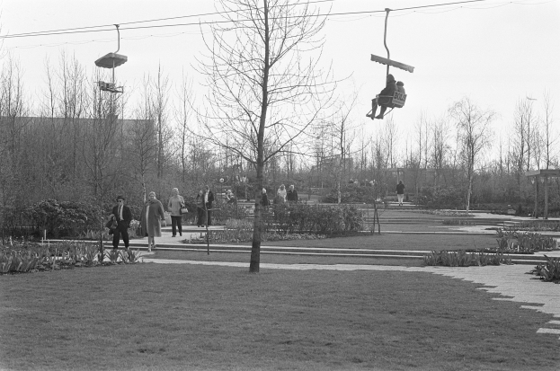 Schwarzes und weißes Foto von Menschen, die eine Skilift in einem Park mit Bäumen, Pflanzen, Gras, einem Weg und einem klaren Himmel im Hintergrund fahren.