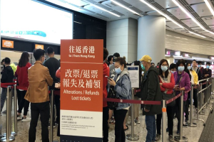 Maskierte Menschen stehen in einer Schlange am Flughafen, getrennt durch Absperrpoller mit Bändern, mit einem Schild im Vordergrund, auf dem "Hong Kong Airport Refunds Due to Covid-19" steht, und Säulen und Deckenleuchten im Hintergrund.