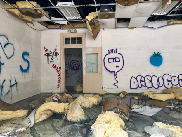 Ein Raum mit Graffiti an den Wänden, eine Tür im Hintergrund, Deckenstangen und Lampen sowie diverse Gegenstände auf dem Boden verstreut.