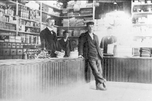 Drei Männer stehen an einem Tresen in einem Geschäft, umgeben von verschiedenen Gegenständen, mit einem Bücherregal und Gegenständen im Hintergrund, scheinbar mitten in einem Einbruch.