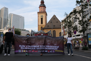 Eine Gruppe maskierter Menschen marschiert auf einer Straße und hält ein Banner hoch, mit einem geparkten Auto auf der linken Seite, Gebäuden, Bäumen, Schildern, Pfosten und einem Uhrenturm im Hintergrund unter einem klaren blauen Himmel.