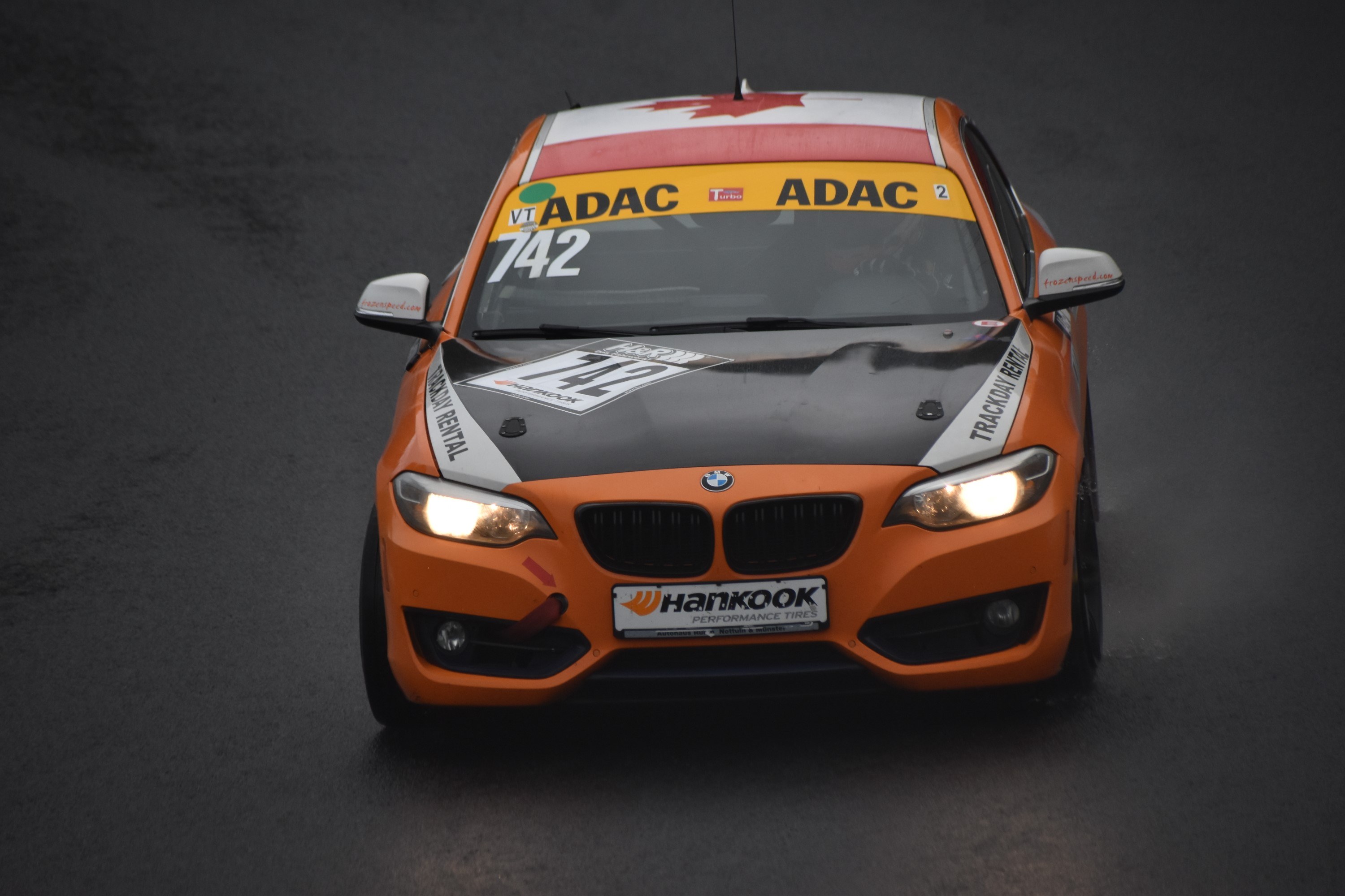 Oranges BMW Rennfahrzeug mit Text und Zahlen auf einer Strecke.