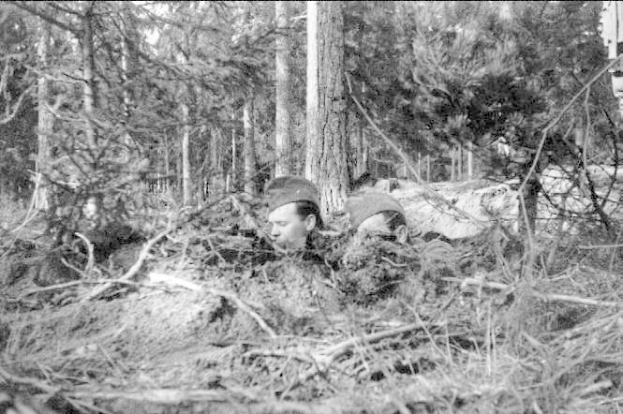 Zwei Männer mit Mützen stehen in einem bewaldeten Gebiet, mit dem Text "Deutsche Soldaten im Wald" am unteren Rand des Schwarz-Weiß-Fotos.