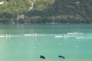 Eine Gruppe von Menschen, die auf einem Gewässer umgeben von Bäumen und einem Hügel rudern, mit Kleiderständern am Boden, die auf einen Ruderwettbewerb hinweisen.