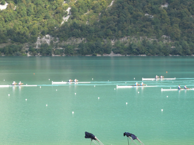 Eine Gruppe von Menschen, die auf einem Gewässer umgeben von Bäumen und einem Hügel rudern, mit Kleiderständern am Boden, die auf einen Ruderwettbewerb hinweisen.