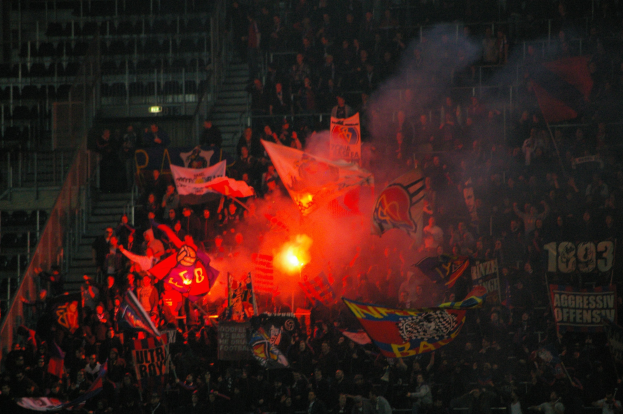 Eine große Menschenmenge in einem Stadion mit Fahnen, Bannern und einem zentralen Feuer, umgeben von Stufen und Geländern, unter Stadionbeleuchtung.