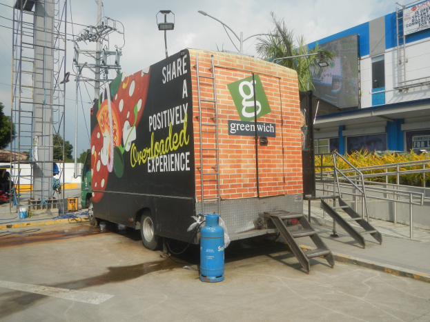 Ein Lkw vor einem Gebäude mit einer Tafel, auf der "Teilen Sie ein positiv überladenes Erlebnis" steht, mit Stufen, einem Zylinder auf dem Boden, Geländern, Strommasten, Laternen, Bäumen und einem bewölkten Himmel.
