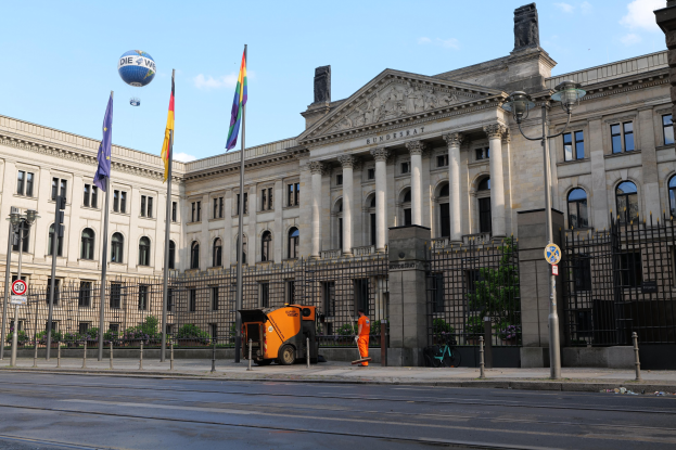 Großes Gebäude mit vielen Fenstern, als Bundestag in Berlin identifiziert, umgeben von Straßeninfrastruktur, Fahrzeugen, Fußgängern und Bäumen unter einem bewölkten Himmel.