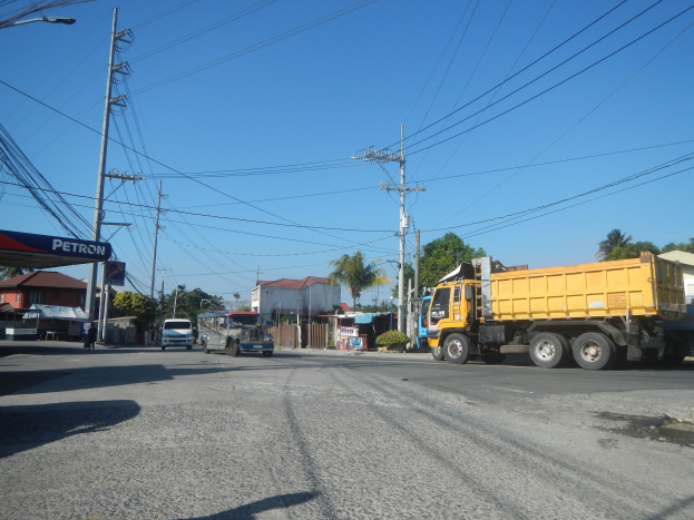 Ein gelber Müllwagen fährt auf einer Straße mit Strommasten, Kabeln, Gebäuden und Bäumen im Hintergrund, mit einer Tankstelle auf der linken Seite und Himmel oben.