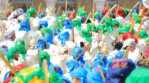 Eine große Gruppe von Menschen in farbenfrohen Kostümen, die eine Straße entlang marschieren und jeweils einen Stock halten, während im Hintergrund ein Gebäude zu sehen ist.