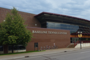 Das Baseline Tennis Center in St. Louis, Missouri, ein großes Gebäude mit Glasfenstern und einem Namen auf dem Schild, mit Treppen zum Eingang, Fahrrädern davor, einem Baum links und einem bewölkten Himmel im Hintergrund.