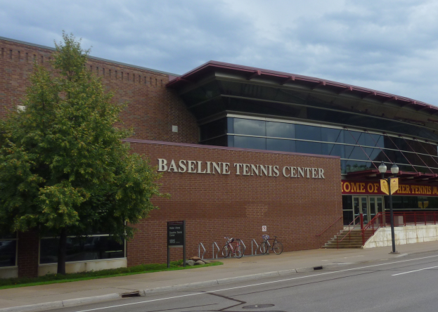 Das Baseline Tennis Center in St. Louis, Missouri, ein großes Gebäude mit Glasfenstern und einem Namen auf dem Schild, mit Treppen zum Eingang, Fahrrädern davor, einem Baum links und einem bewölkten Himmel im Hintergrund.