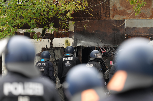 Eine Gruppe von Polizisten in schwarzen Uniformen und Helmen steht vor einem Gebäude, das von einem Maschendrahtzaun umgeben ist. Im Hintergrund sind Bäume zu sehen.