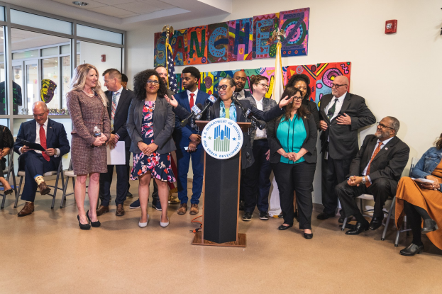 Gruppe von Menschen vor einem Rednerpult mit Mikrofon und einer Tafel mit der Aufschrift 'Sancho-Stadtrat tritt dem Stadtrat bei', mit sitzenden und stehenden Teilnehmern, Fahnen, Feuerlöscher und Deckenleuchten im Hintergrund.