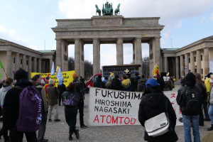 Eine Gruppe von Menschen steht vor dem Brandenburger Tor in Berlin, Deutschland, mit Schildern und Fahnen, auf denen 'Fukushima Hiroshima Nagasaki Atomod' steht, um gegen die Atombombe zu protestieren, mit Fahrzeugen, Bäumen und einem bewölktem Himmel im Hintergrund.