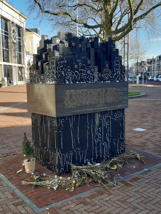 Denkmal in einem Stadtplatz, das den Opfern des Holocaust gewidmet ist, mit einem Baum davor, einem Topf mit einer Pflanze, verstreuten trockenen Blättern und im Hintergrund Gebäude, Fahrzeuge, Masten und Bäume.