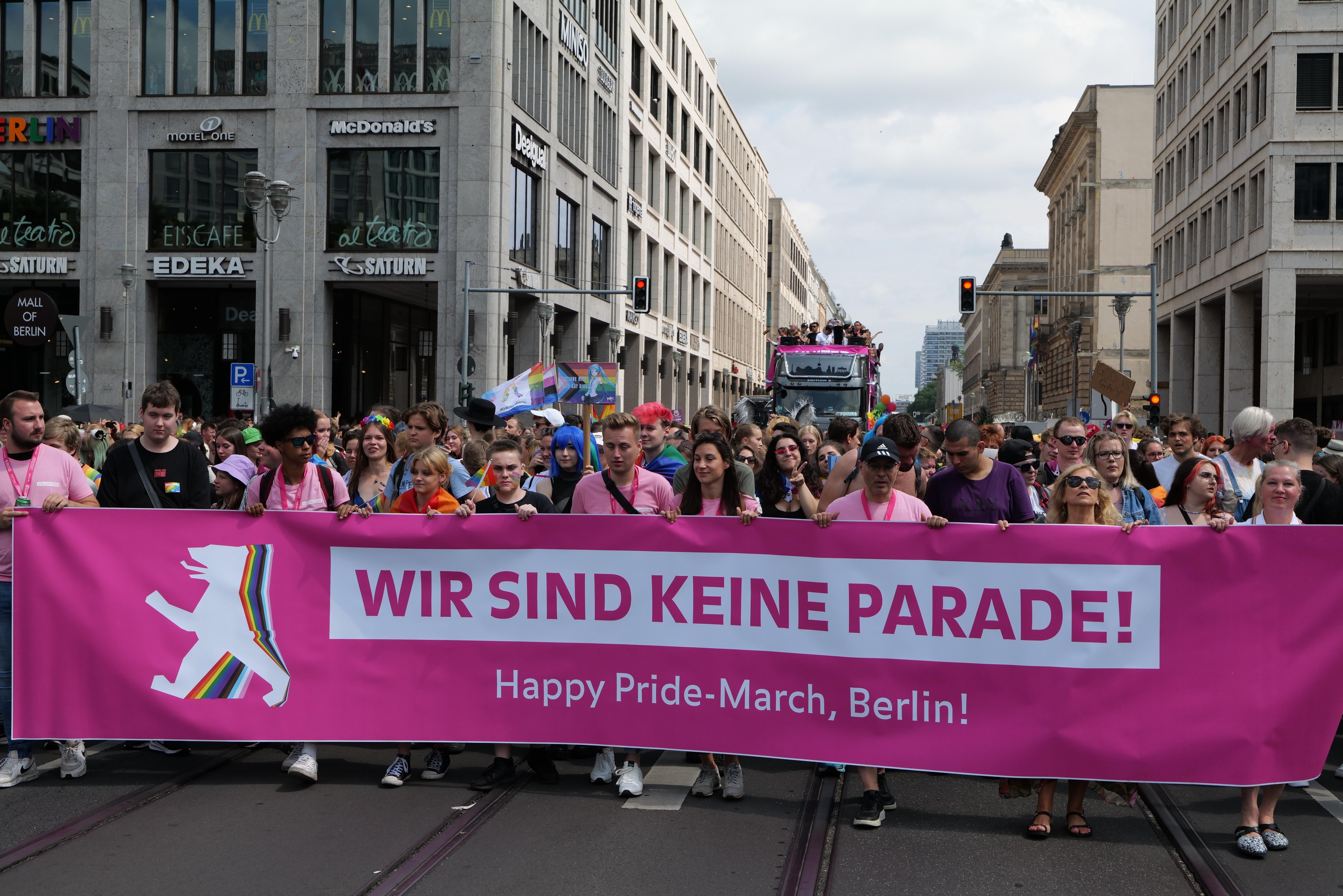 Eine Gruppe von Menschen marschiert eine Berliner Straße entlang und hält eine pinkfarbene 'Happy Pride March'-Fahne hoch, während Gebäude, Laternenpfähle und Verkehrszeichen die Straße säumen.