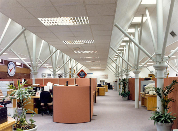 Leerer Büroraum mit ordentlich angeordneten Schreibtischen und Stühlen, Topfpflanzen, tragenden Säulen, einer Wanduhr und Deckenleuchten.