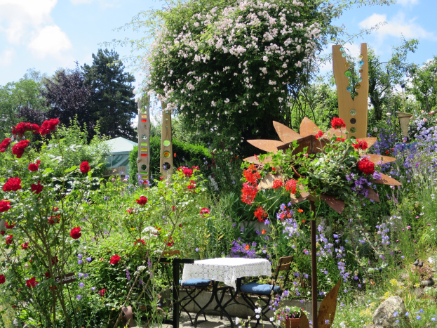 Ein Garten auf der Chelsea Flower Show mit bunten Blumen und Pflanzen, einem Tisch mit einer Decke und Stühlen und Bäumen im Hintergrund unter einem bewölkten Himmel.