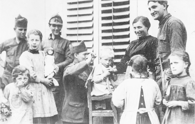 Schwarz-weiß-Fotografie einer Gruppe von Flüchtlingen, die vor einem Gebäude stehen, mit einer Frau auf einem Stuhl in der Mitte, einige halten Kinder, und ein Fenster ist im Hintergrund zu sehen.