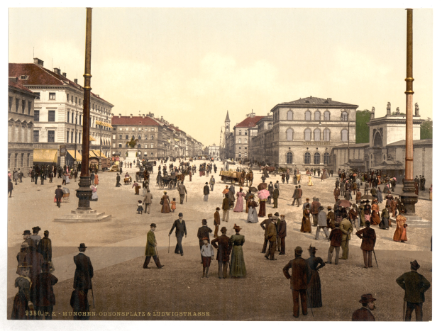 Schwarz-weiß-Foto einer belebten Münchner Straße in den frühen 1900er Jahren mit Passanten, einem Pferdewagen, Gebäuden, Bäumen und einer Unterschrift am unteren Rand.
