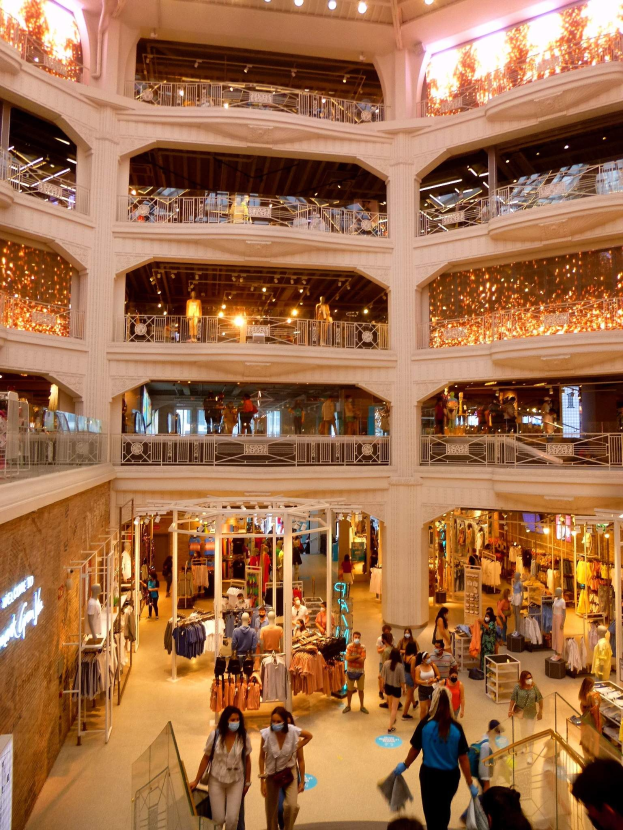 Großer Atrium eines belebten Einkaufszentrums mit Menschen, die Taschen tragen und Kleiderauslagen auf Schaufensterpuppen in der Nähe von Geländern und beleuchteten Wänden mit Texttafeln betrachten.