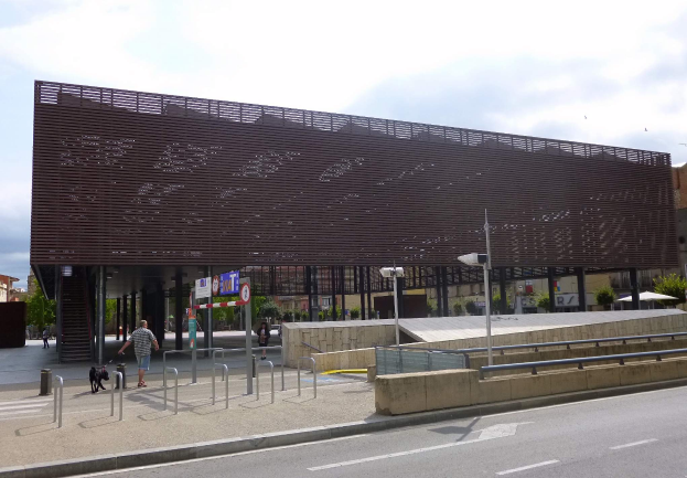Ein großes Gebäude mit vielen Fenstern dient als neuer Eingang zur City, umgeben von Straßenlaternen, Fahrzeugen, Fußgängern, Sonnenschirmen, Bäumen und einem bewölkten Himmel.