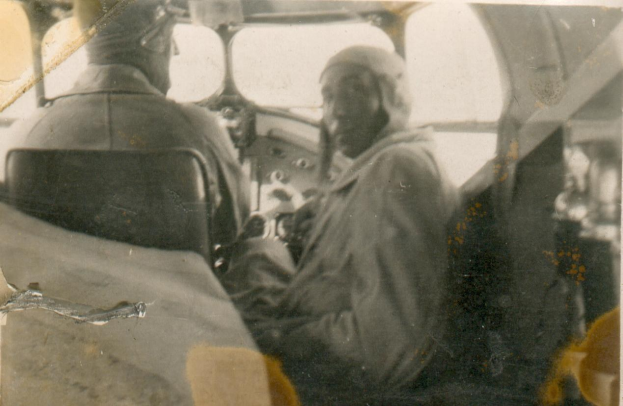 Schwarzes und weißes Foto von zwei Männern in einem Cockpit; einer sitzt im Pilotensitz und trägt eine Uniform, der andere steht neben ihm und hat einen ernsten Gesichtsausdruck.