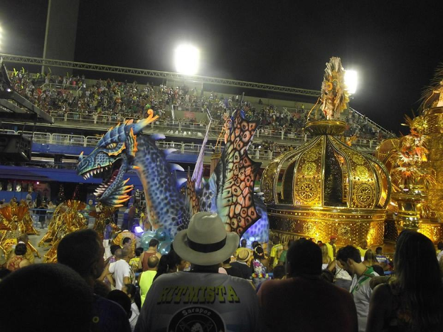 Eine große Menschenmenge umringt einen bunt geschmückten und hell erleuchteten Karnevalswagen, im Hintergrund ist ein Stadion und der Himmel zu sehen.