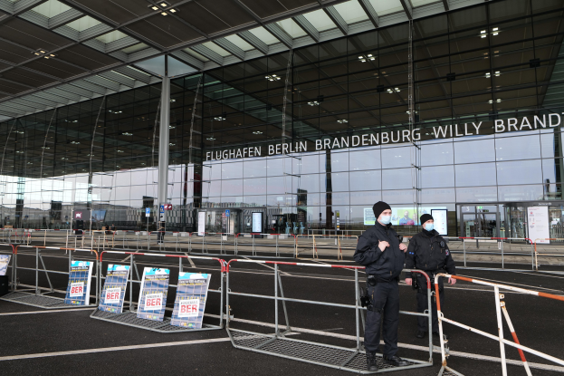 Zwei Polizisten in schwarzen Uniformen stehen vor einem großen Glasgebäude mit Text darauf, mit Absperrungen und Schildern im Vordergrund und Menschen, Polen und Lichter im Hintergrund.