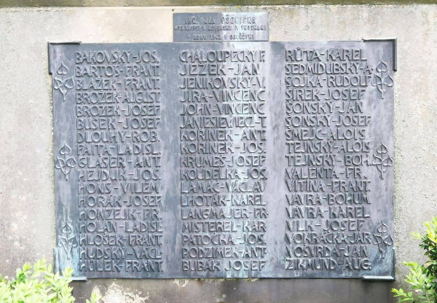 Ein Steinplakette an einer Wand mit eingravierter Schrift, umgeben von Pflanzen am Boden, wahrscheinlich ein Holocaust-Mahnmal.