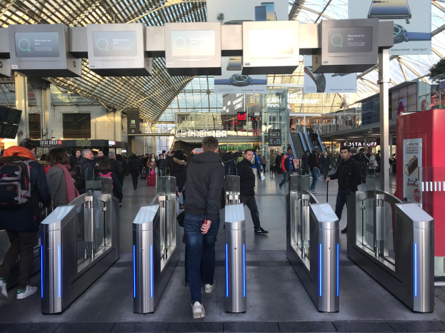 Eine Gruppe von Menschen geht durch einen belebten Bahnhof mit Metalldetektoren, Texttafeln, Säulen, Rolltreppen und Drehkreuzen, unter einer sichtbaren Decke.