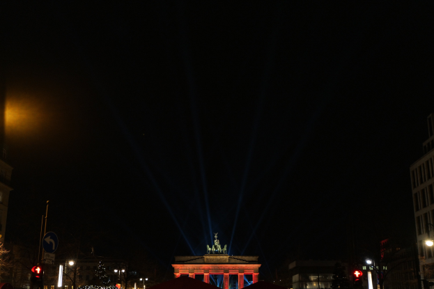 Brandenburger Tor bei Nacht, beleuchtet von Straßenlaternen, mit Gebäuden, Bäumen, Pfählen, Verkehrszeichen, Schildern und Zelten drumherum vor dunklem Himmel.